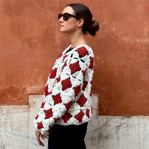Op de afbeelding: Een wit en rood gehaakt vest met een geometrisch patroon. Het vest wordt gedragen over een zwarte top en gecombineerd met een zwarte broek. De achtergrond is een getextureerde, roodbruine muur.