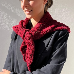 Puede incluir: Bufanda de ganchillo rojo oscuro sobre una blazer negra. La bufanda tiene un diseño calado y está envuelta alrededor del cuello. La blazer tiene un corte clásico y está abotonada. El fondo es una pared lisa de color blanquecino.
