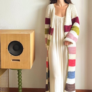 May include: A long, open-front crochet cardigan with colorful horizontal stripes in shades of cream, burgundy, green, pink, red, blue, and brown. The cardigan is worn over a white dress. A wooden speaker sits on a green stand.