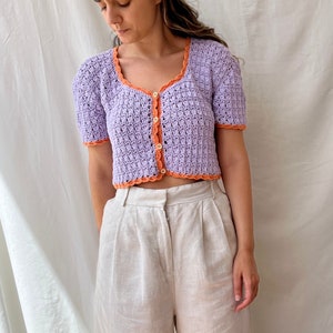 May include: A lavender crochet top with a light orange trim and a single button closure. The top has short sleeves and a cropped length.