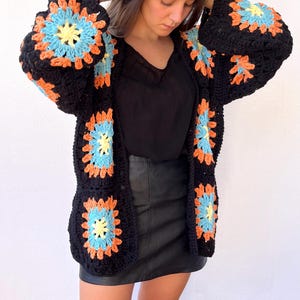 Pode incluir: Cardigã preto de crochê com padrões florais coloridos em laranja, azul e amarelo. O suéter aberto apresenta mangas compridas e um design único feito à mão. Uma peça de roupa elegante e confortável para sobrepor.