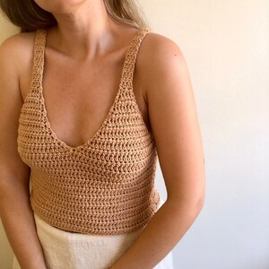 May include: A crocheted tank top with a v-neck and thin straps. The top is made of a light brown yarn.