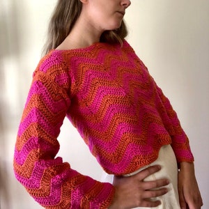 May include: A pink and orange crocheted sweater with a chevron pattern. The sweater has long sleeves and a cropped length.