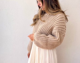 Könnte beinhalten: Ein beiger Strickpullover mit langen Ärmeln und einer verkürzten Länge. Der Pullover hat eine lockere Passform und ein strukturiertes Strickmuster.