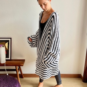 May include: A long, open-front crochet cardigan in a bold black and white striped pattern. The handmade garment features a relaxed fit with wide sleeves and a knee-length hem. The cardigan is worn over a black top and leggings.