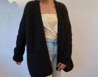 May include: A black crocheted cardigan with a long, loose fit. The cardigan is open in the front and has a simple, classic design.