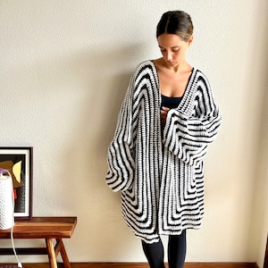 May include: A black and white crochet cardigan with a geometric pattern. The long-sleeved cardigan is knee-length and has a V-neck. The open-front design features a bold, eye-catching pattern.