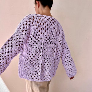 Puede incluir: Una chaqueta de punto de ganchillo de color lavanda con un patrón de punto cuadrado. La chaqueta tiene mangas largas y un corte holgado.