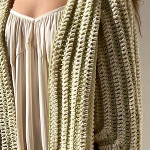 May include: A cream-colored dress is partially visible beneath a green and cream crochet cardigan. The cardigan has a ribbed texture and a loose, open design. The color palette is soft and neutral, with a focus on natural tones and textures.