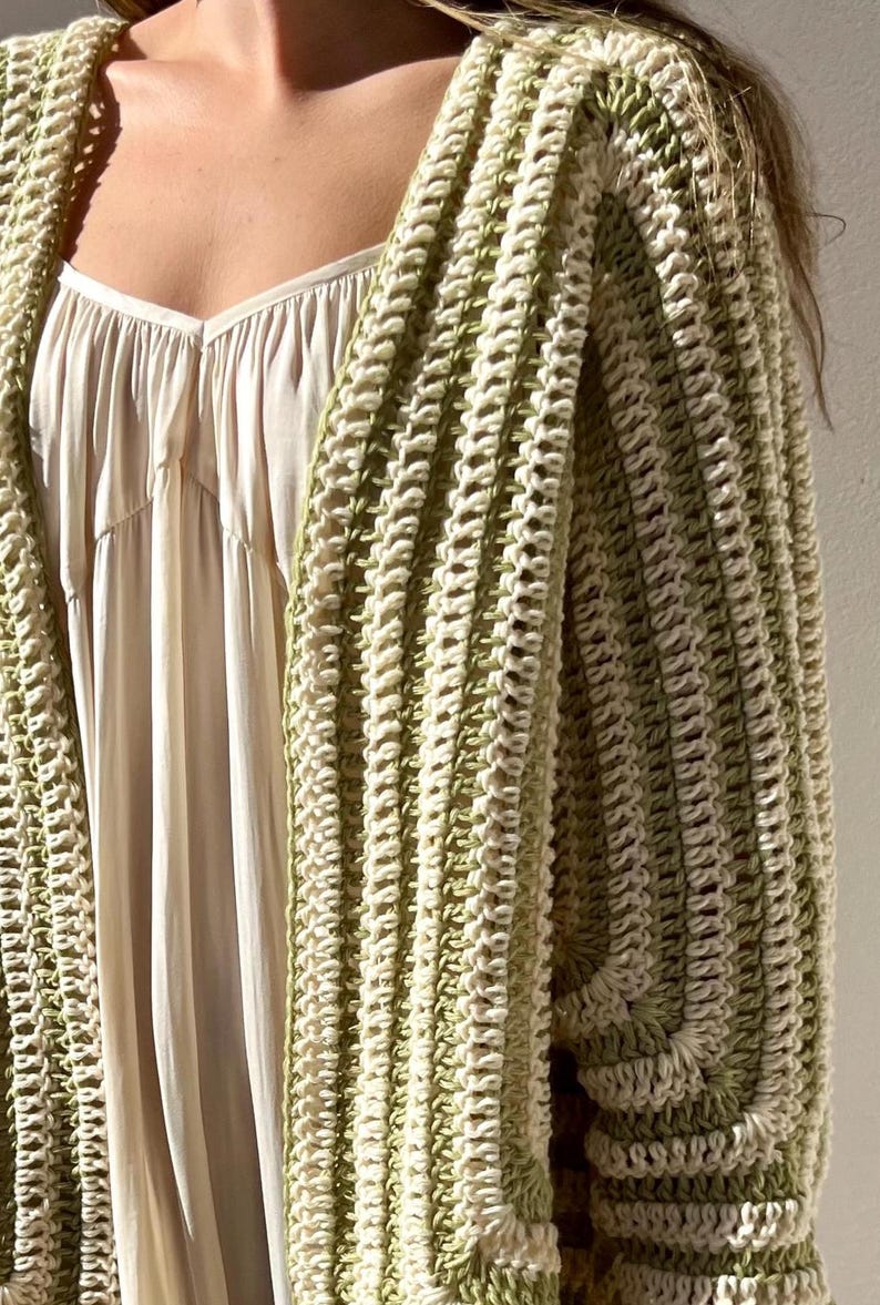 May include: A cream-colored dress is partially visible beneath a green and cream crochet cardigan. The cardigan has a ribbed texture and a loose, open design. The color palette is soft and neutral, with a focus on natural tones and textures.