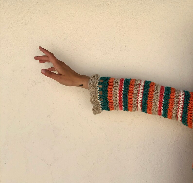 May include: A close-up of a striped crochet sleeve in orange, green, beige, and pink. The sleeve is partially rolled up, revealing a beige crocheted cuff.