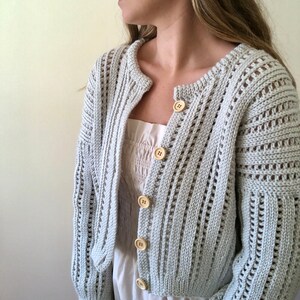 K&ouml;nnte beinhalten: Ein hellgrauer Strickcardigan mit Knopfleiste und leicht verk&uuml;rzter L&auml;nge. Der Cardigan hat ein zartes Lochmuster und ist aus weichem, leichtem Garn gefertigt.