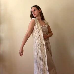 Puede incluir: Un chaleco largo de crochet blanco con un delicado patr&oacute;n de punto abierto. El chaleco es sin mangas y tiene un escote en V. Se lleva sobre un vestido marr&oacute;n claro.