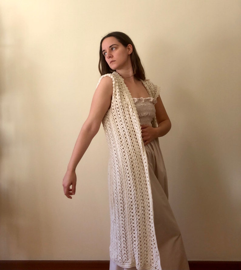 Puede incluir: Un chaleco largo de crochet blanco con un delicado patr&oacute;n de punto abierto. El chaleco es sin mangas y tiene un escote en V. Se lleva sobre un vestido marr&oacute;n claro.