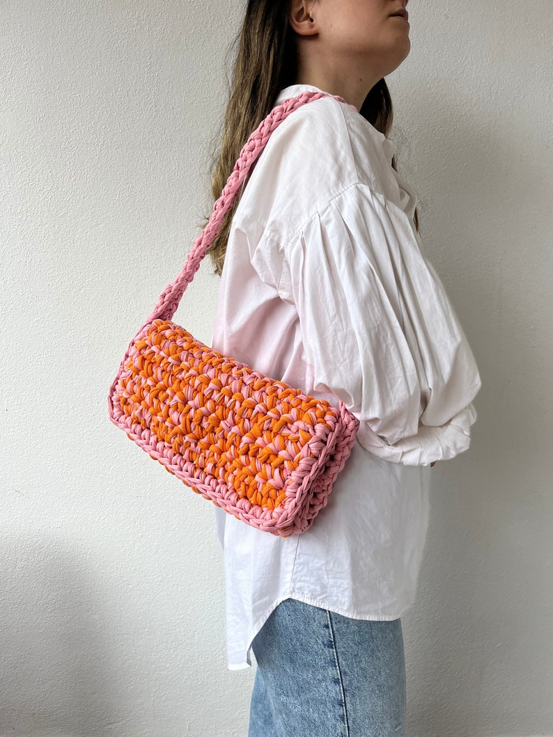 May include: A pink and orange crocheted shoulder bag. The bag is made with a thick yarn and has a simple, rectangular shape.