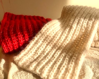 May include: Two knitted scarves, one white and one red, are displayed on a textured beige surface. The white scarf has a brown leather tag with the word "silvcrim" printed on it.