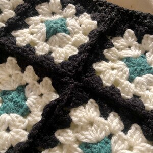 May include: Close-up of a crocheted blanket with a black and white pattern and teal accents. The blanket is made of yarn and has a textured surface.