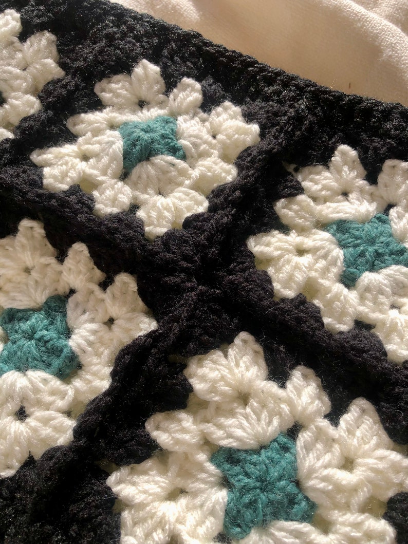 May include: Close-up of a crocheted blanket with a black and white pattern and teal accents. The blanket is made of yarn and has a textured surface.
