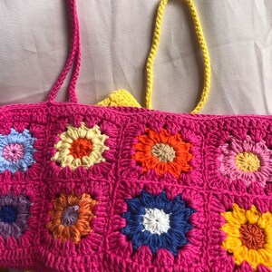 K&ouml;nnte beinhalten: Eine handgefertigte H&auml;keltasche mit einem lebendigen Blumenmuster. Die Tasche hat eine pinkfarbene Basis mit bunten Blumenquadraten in Blau-, Gelb-, Orange- und Rosat&ouml;nen. Die Tasche hat rosa Tr&auml;ger und ein gelbes Futter.