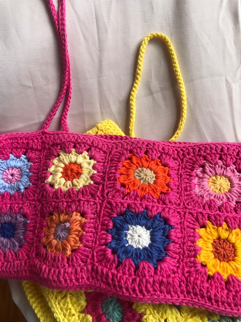 K&ouml;nnte beinhalten: Eine handgefertigte H&auml;keltasche mit einem lebendigen Blumenmuster. Die Tasche hat eine pinkfarbene Basis mit bunten Blumenquadraten in Blau-, Gelb-, Orange- und Rosat&ouml;nen. Die Tasche hat rosa Tr&auml;ger und ein gelbes Futter.
