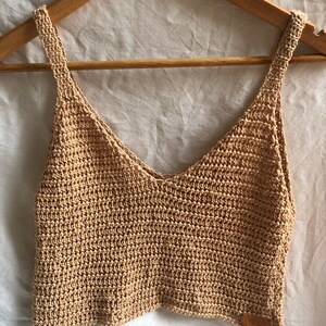 May include: A beige crochet tank top with a v-neck and thin straps. The top is made of a lightweight, breathable material and has a relaxed fit.