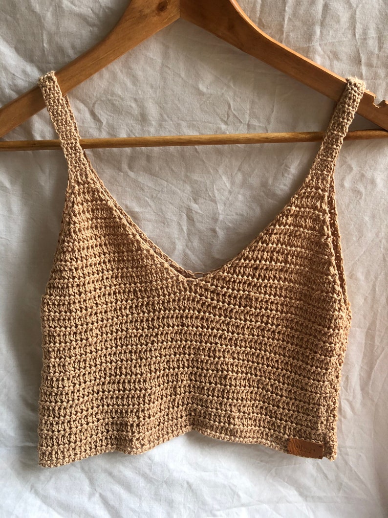 May include: A beige crochet tank top with a v-neck and thin straps. The top is made of a lightweight, breathable material and has a relaxed fit.