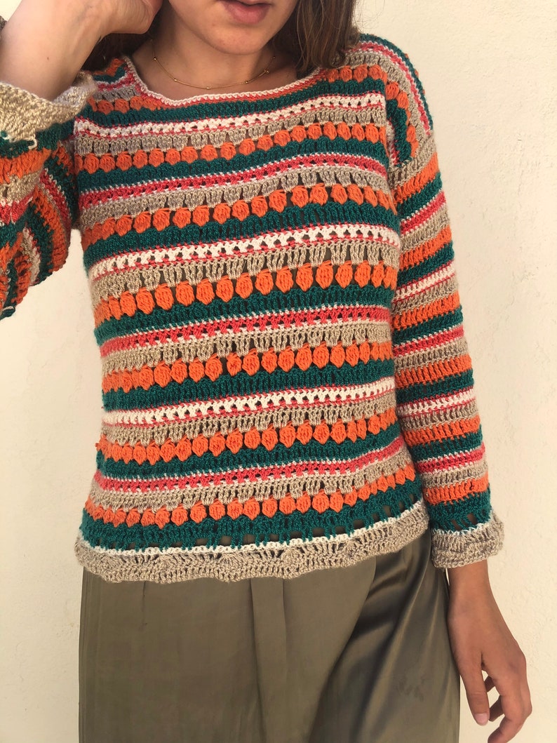 May include: A striped crochet sweater with long sleeves in shades of orange, green, beige, and white. The sweater has a loose fit and a round neckline.