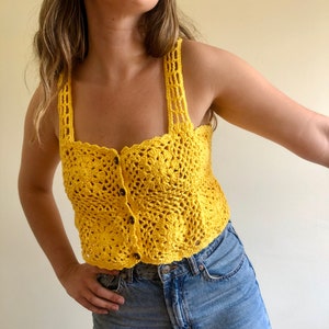 Puede incluir: Un top de crochet amarillo con cierre de botón. El top tiene un escote cuadrado y tirantes finos.
