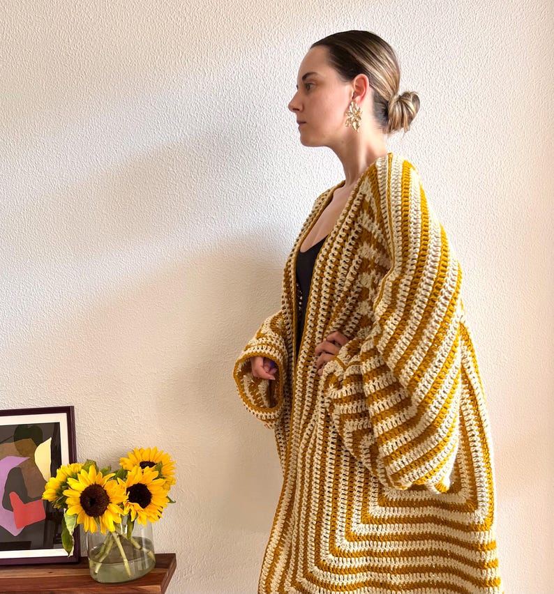 May include: A mustard yellow and cream crochet cardigan with a geometric pattern. The long-sleeved, open-front cardigan is knee-length and has a relaxed fit. The cardigan is handmade and appears to be a cozy, bohemian style.