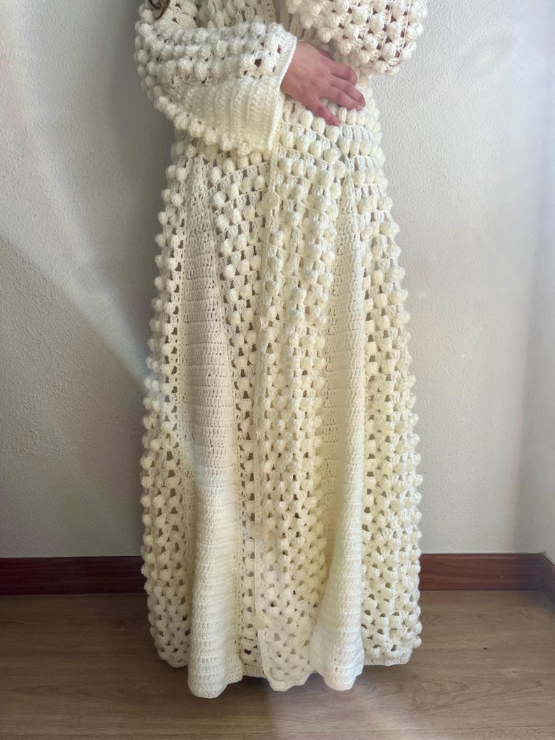 May include: A white crocheted maxi dress with a textured, bobble stitch pattern. The dress has a long, flowing skirt and a fitted bodice.