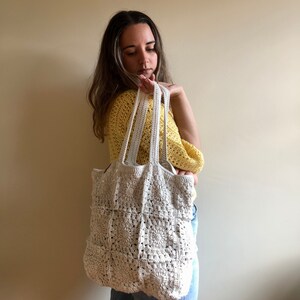 May include: A white crocheted tote bag with a square pattern. The bag has two straps and is being held by a person wearing a yellow shirt and blue jeans.
