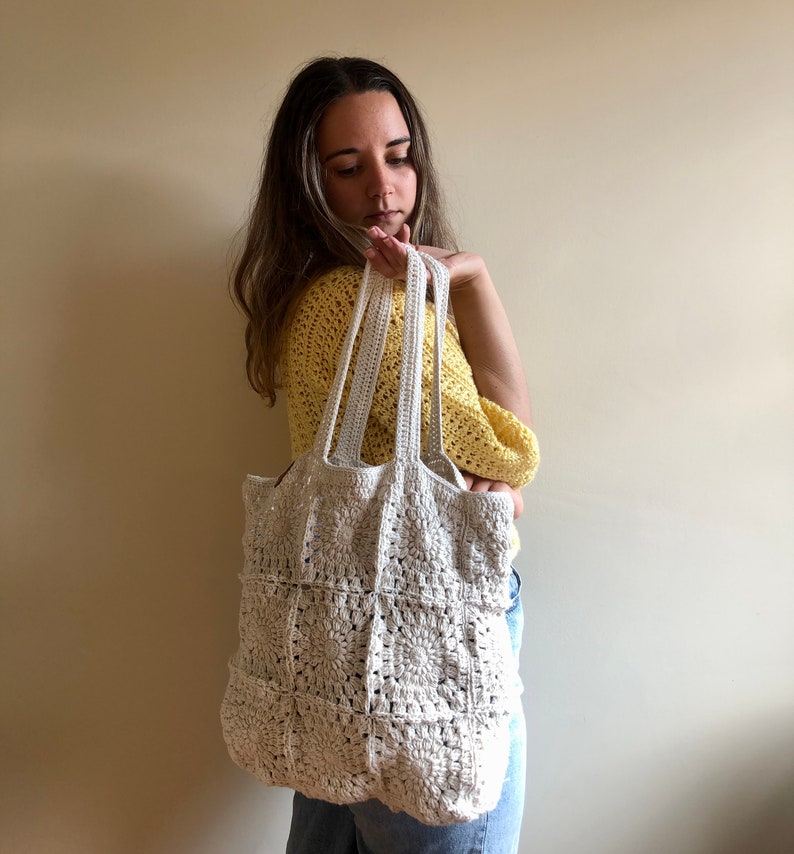 May include: A white crocheted tote bag with a square pattern. The bag has two straps and is being held by a person wearing a yellow shirt and blue jeans.