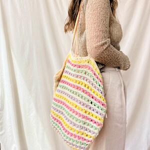 May include: A colorful striped crochet tote bag with a light green, yellow, pink, and purple color scheme. The bag is being held by a person wearing a beige sweater.