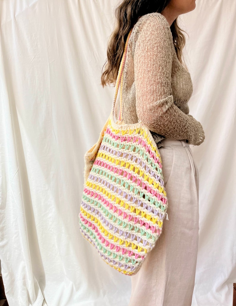 May include: A colorful striped crochet tote bag with a light green, yellow, pink, and purple color scheme. The bag is being held by a person wearing a beige sweater.