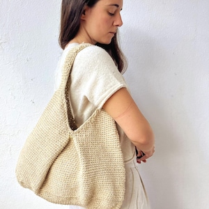 Puede incluir: Un bolso de hombro beige tejido a mano. El bolso tiene una forma redondeada con una correa ancha y un patrón de puntada texturizada. El bolso está hecho de un hilo de color natural, y el diseño general es simple y funcional.