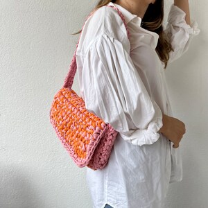 May include: A pink and orange crocheted shoulder bag with a long strap.