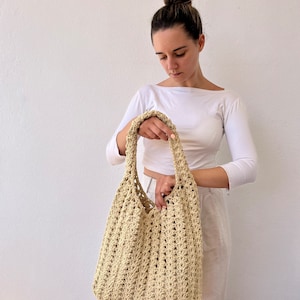 K&ouml;nnte beinhalten: Eine beige geh&auml;kelte Tragetasche mit einer breiten, offenen Oberseite und einem einzigen Trageriemen. Die Tasche ist aus einem zarten, offen gewebten Muster gefertigt.
