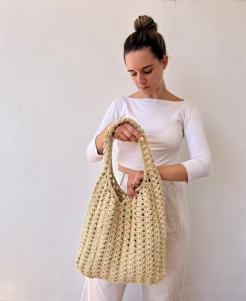 K&ouml;nnte beinhalten: Eine beige geh&auml;kelte Tragetasche mit einer breiten, offenen Oberseite und einem einzigen Trageriemen. Die Tasche ist aus einem zarten, offen gewebten Muster gefertigt.