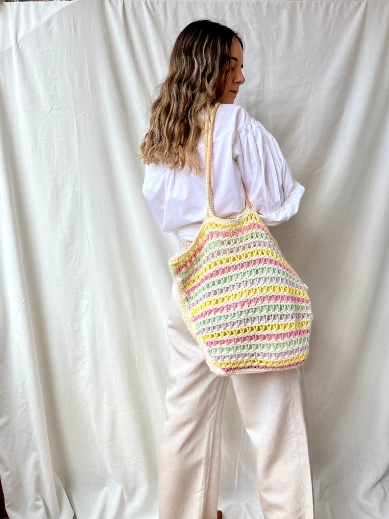 May include: A large, colorful crochet tote bag with a striped pattern in shades of pink, yellow, green, and white. The bag is worn over the shoulder of a person wearing a white shirt and beige pants.