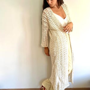 Puede incluir: Un cárdigan largo de ganchillo color crema con un patrón geométrico de punto abierto, mangas largas y una silueta recta hasta el suelo. La prenda se lleva sobre una camiseta blanca y pantalones claros, combinada con sandalias rojas de tacón.