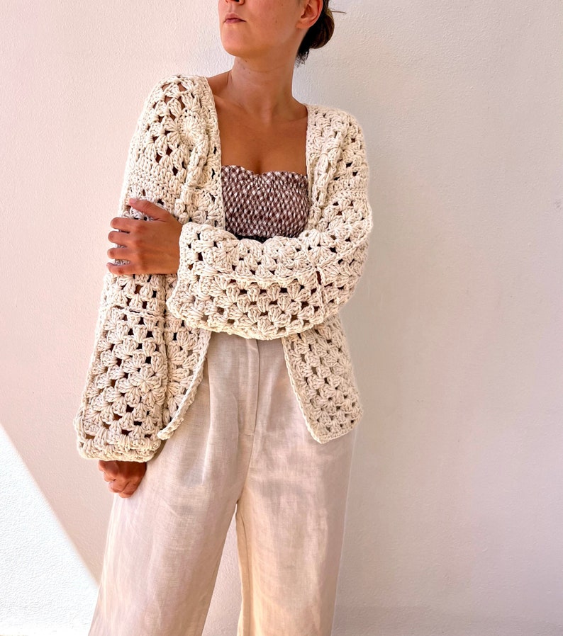 May include: A cream-colored crocheted cardigan with a square stitch pattern. The cardigan has a relaxed fit and long sleeves.