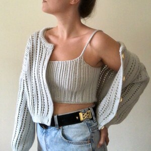 K&ouml;nnte beinhalten: Ein hellgrauer H&auml;kelcardigan und ein passendes Crop-Top mit einem schwarzen G&uuml;rtel und einer goldenen Schnalle.
