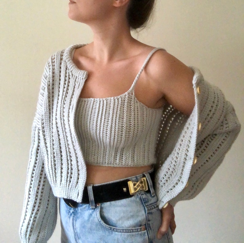 K&ouml;nnte beinhalten: Ein hellgrauer H&auml;kelcardigan und ein passendes Crop-Top mit einem schwarzen G&uuml;rtel und einer goldenen Schnalle.