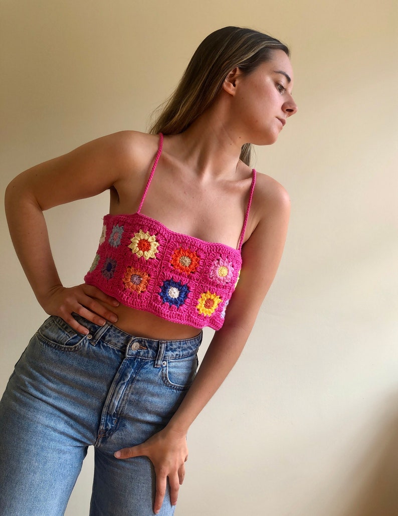 May include: A pink crochet top with colorful flower designs. The top has thin straps and a cropped fit.