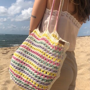 May include: A crocheted tote bag with a colorful striped pattern in pink, yellow, green, and purple. The bag has a white crocheted strap and is being carried by a person on a beach.