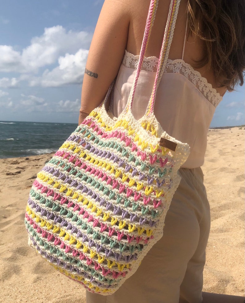 May include: A crocheted tote bag with a colorful striped pattern in pink, yellow, green, and purple. The bag has a white crocheted strap and is being carried by a person on a beach.