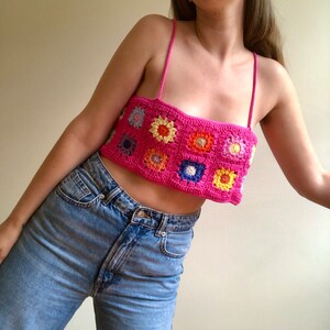 K&ouml;nnte beinhalten: Ein leuchtend pinkes H&auml;keltop mit bunten Blumenquadraten. Das Top hat d&uuml;nne Tr&auml;ger und wird mit hellblauen Jeans getragen. Ein handgefertigtes Kleidungsst&uuml;ck.
