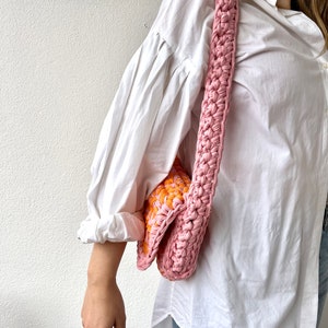 May include: A pink and orange crocheted shoulder bag with a long strap. The bag is worn over the shoulder of a person wearing a white shirt and blue jeans.