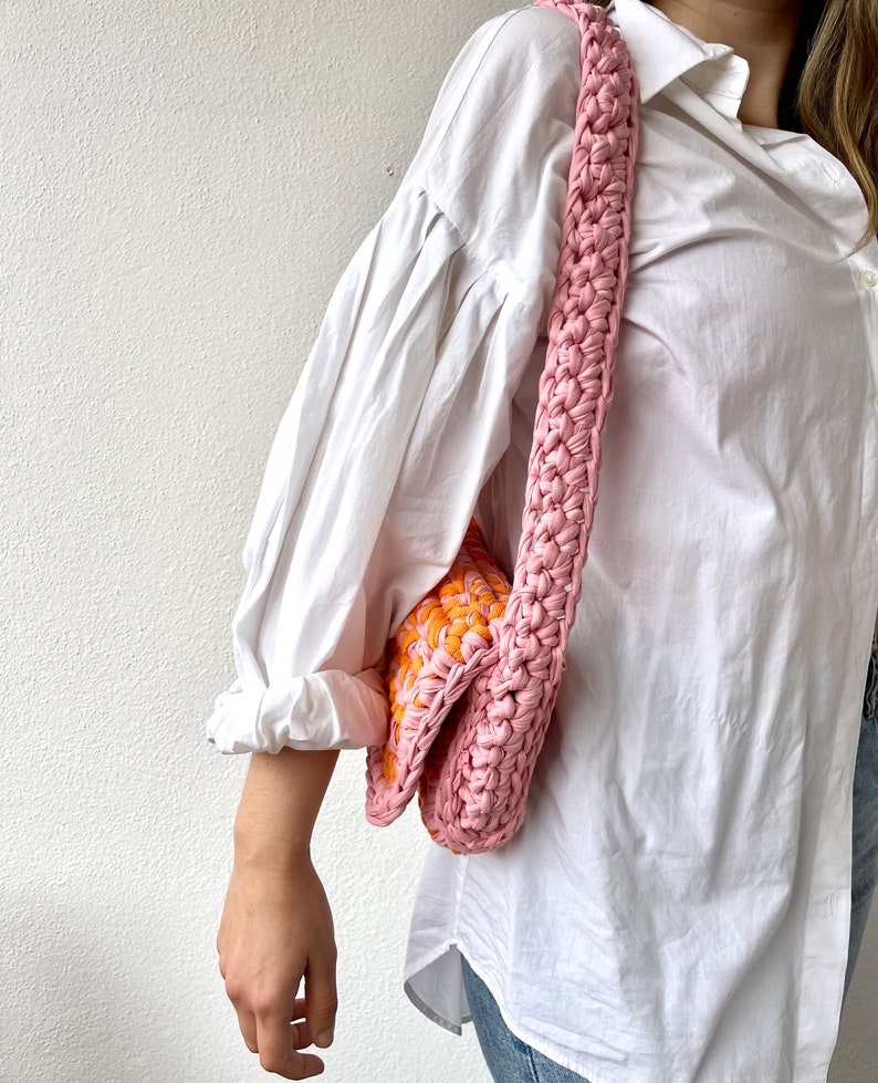 May include: A pink and orange crocheted shoulder bag with a long strap. The bag is worn over the shoulder of a person wearing a white shirt and blue jeans.