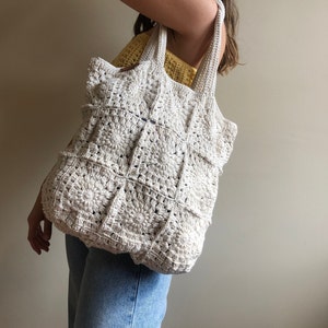 May include: A white crocheted tote bag with a square pattern. The bag has a wide, flat base and two straps.